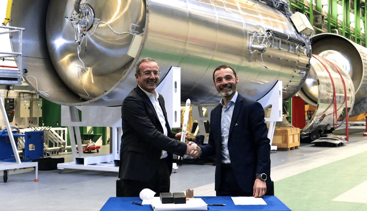 Two men in business attire shake hands in a large industrial hangar in front of massive metallic rocket stages, with one man holding a small model rocket.