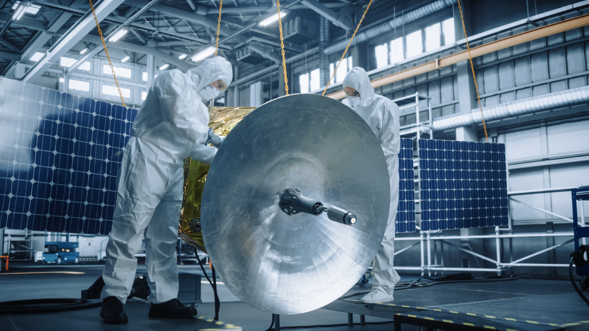 Engineers in cleanroom suits assembling a software-defined satellite with solar panels and a large dish in an MDA factory.