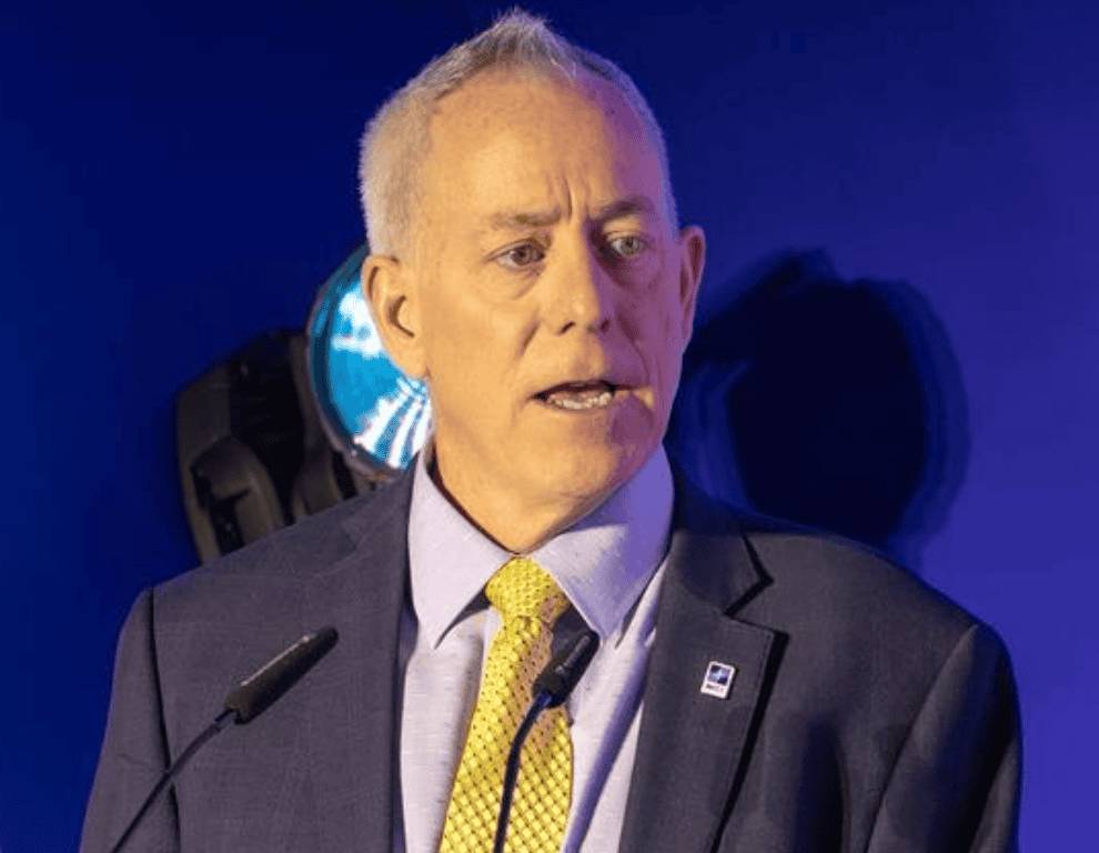 John Blythe, chief of the NATO Integrated Mission Service Center, speaks into microphones while wearing a suit and a yellow tie against a blue background.
