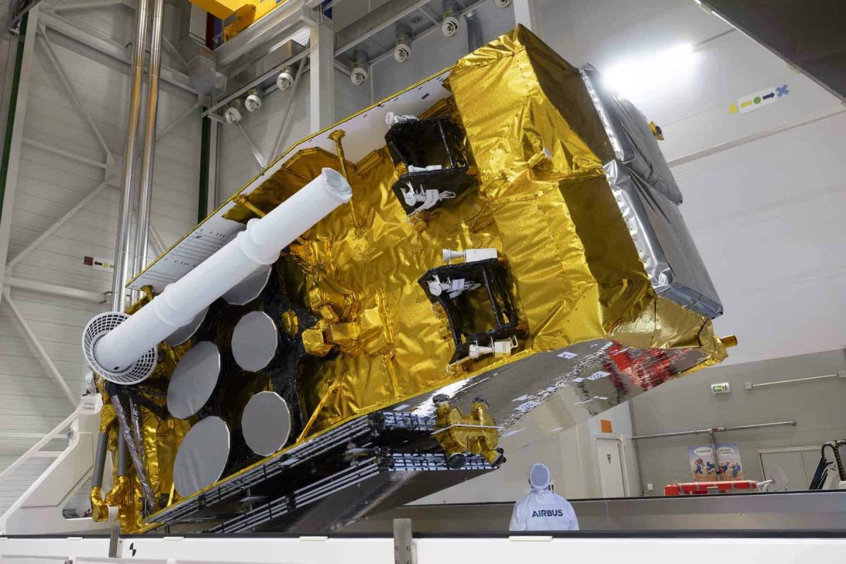 The massive, gold-wrapped SpainSat NG 2 satellite is tilted on a mounting rig in a clean room facility, towering over a worker wearing a white Airbus protective suit.