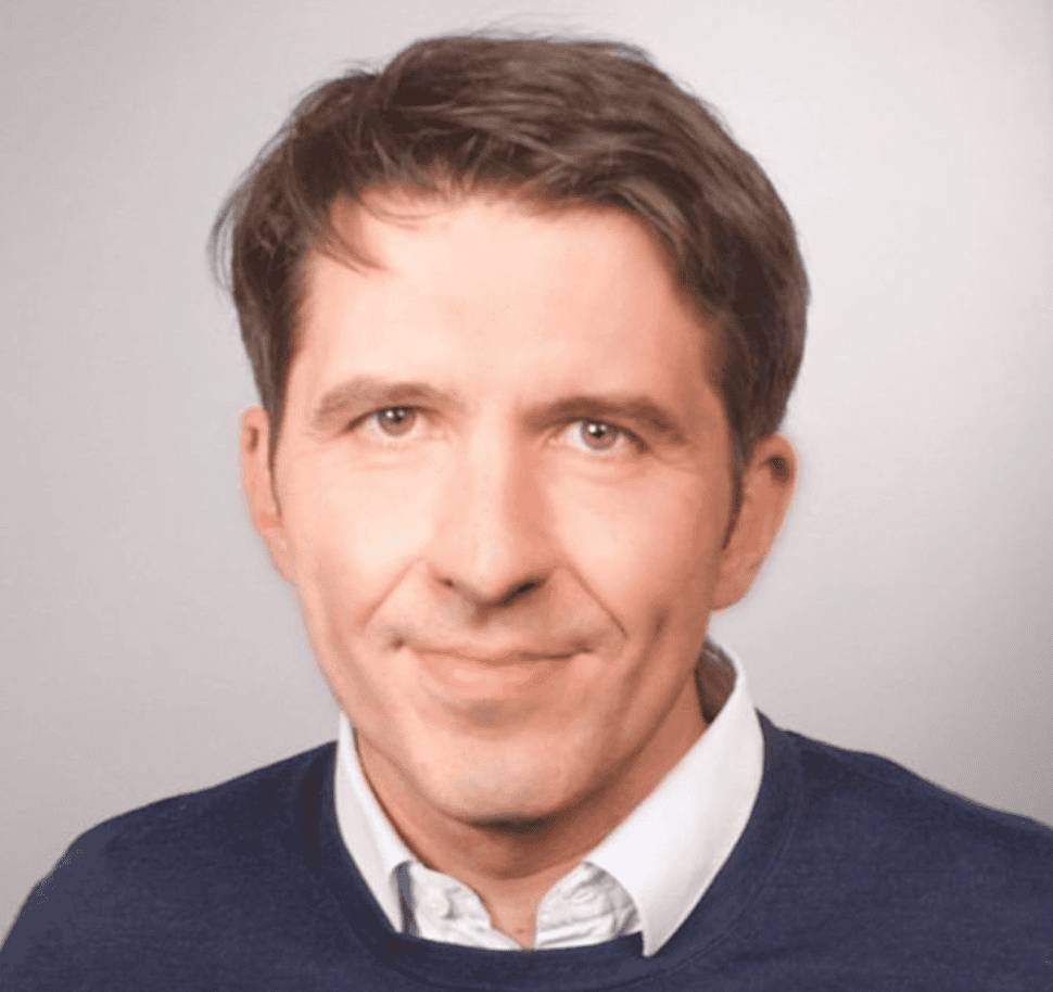A headshot of Charles Delfieux, a man with brown hair wearing a white collared shirt under a dark blue sweater, set against a plain light gray background.