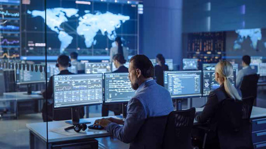 A group of operators sit at rows of computer terminals in a modern, blue-lit security operations center, facing a large central screen displaying a digital world map.