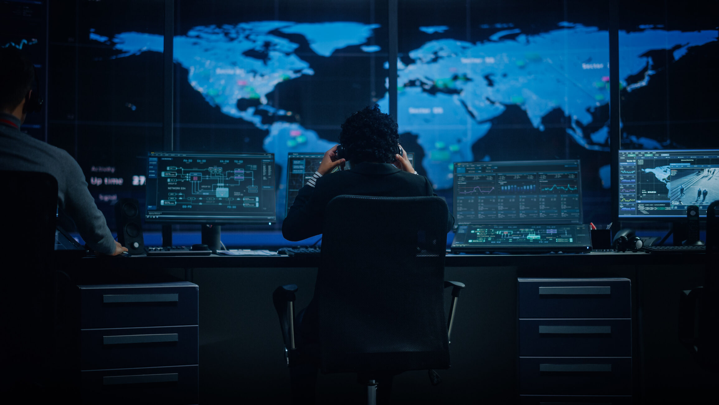 Female analyst sitting in front of dual widescreen monitors displaying network metrics and maps in a darkened room.