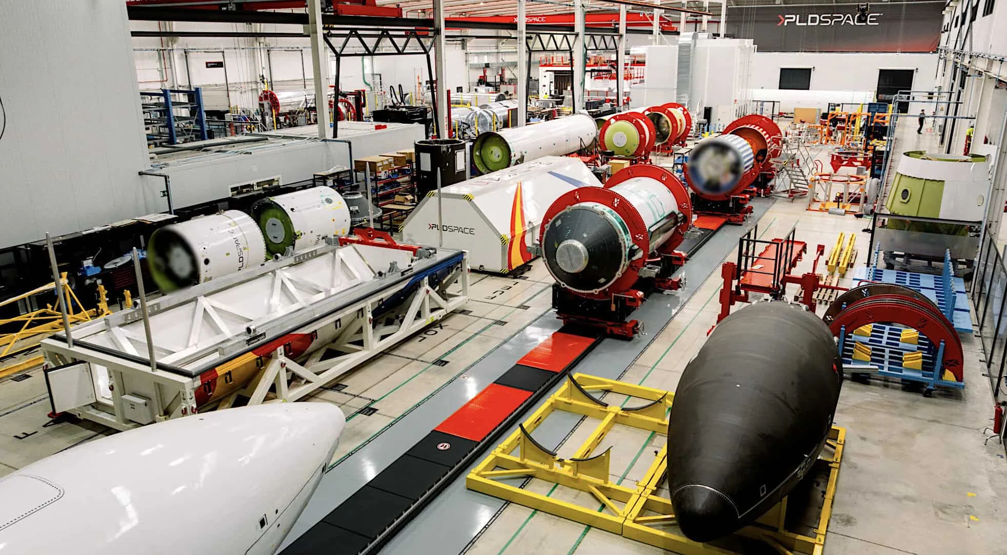 An overhead view inside a PLD Space production facility reveals multiple rocket fuselage sections, fairings, and structural components in various stages of assembly on the factory floor.