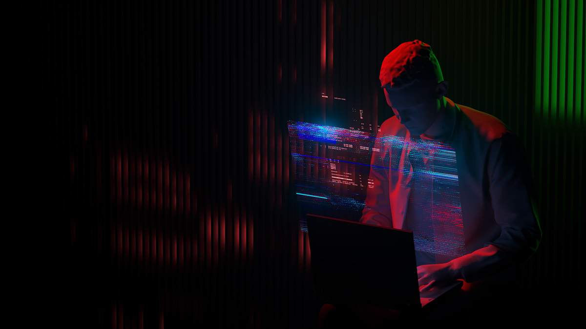 Cybersecurity analyst working on a laptop surrounded by glowing digital code and data telemetry under dramatic red and green ambient lighting.
