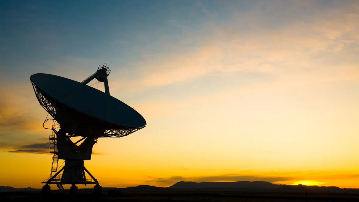 A large radio telescope antenna is silhouetted against a vibrant orange and yellow sunset sky over a distant mountain range.