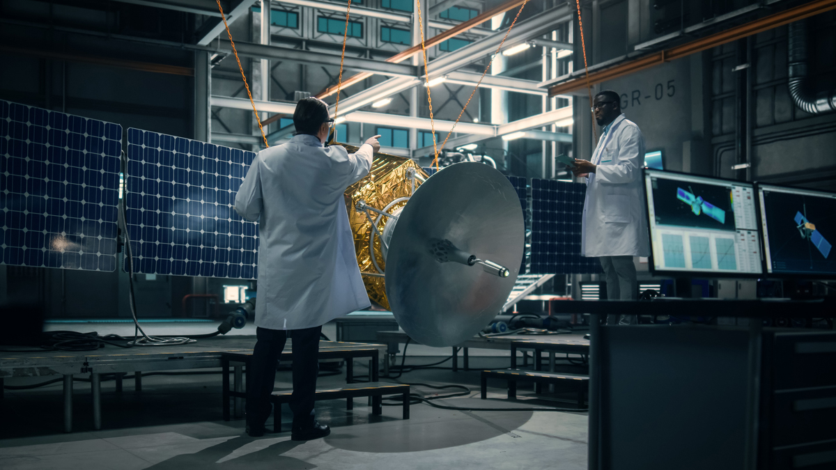 Two aerospace engineers in white lab coats inspecting a satellite and communications dish antenna inside a spacecraft manufacturing facility.