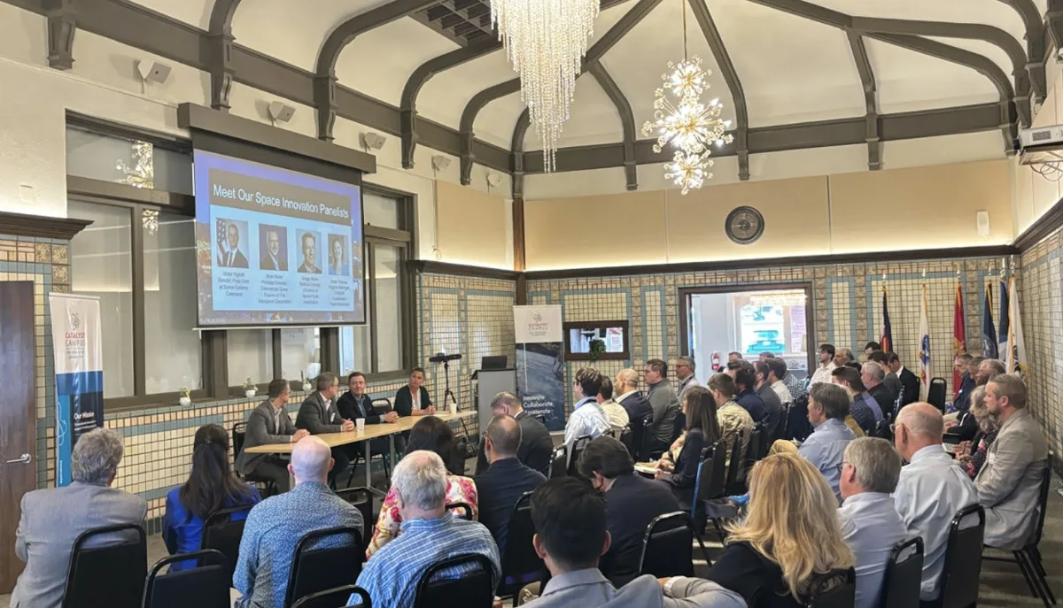 A room full of people attentively listen to a panel of speakers discussing space innovation, with a large screen displaying panelist information and banners indicating the event's association with Catalyst Campus.