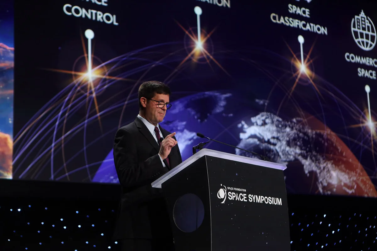 A man is speaking at a podium labeled 'Space Symposium' with a backdrop displaying space-related themes and text, such as 'Space Control' and 'Space Classification'.