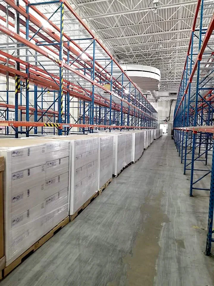 A warehouse interior with rows of tall, empty blue and orange shelving on the right, and a long line of wrapped pallets containing Hughes OneWeb antennas on the left, ready for shipment.