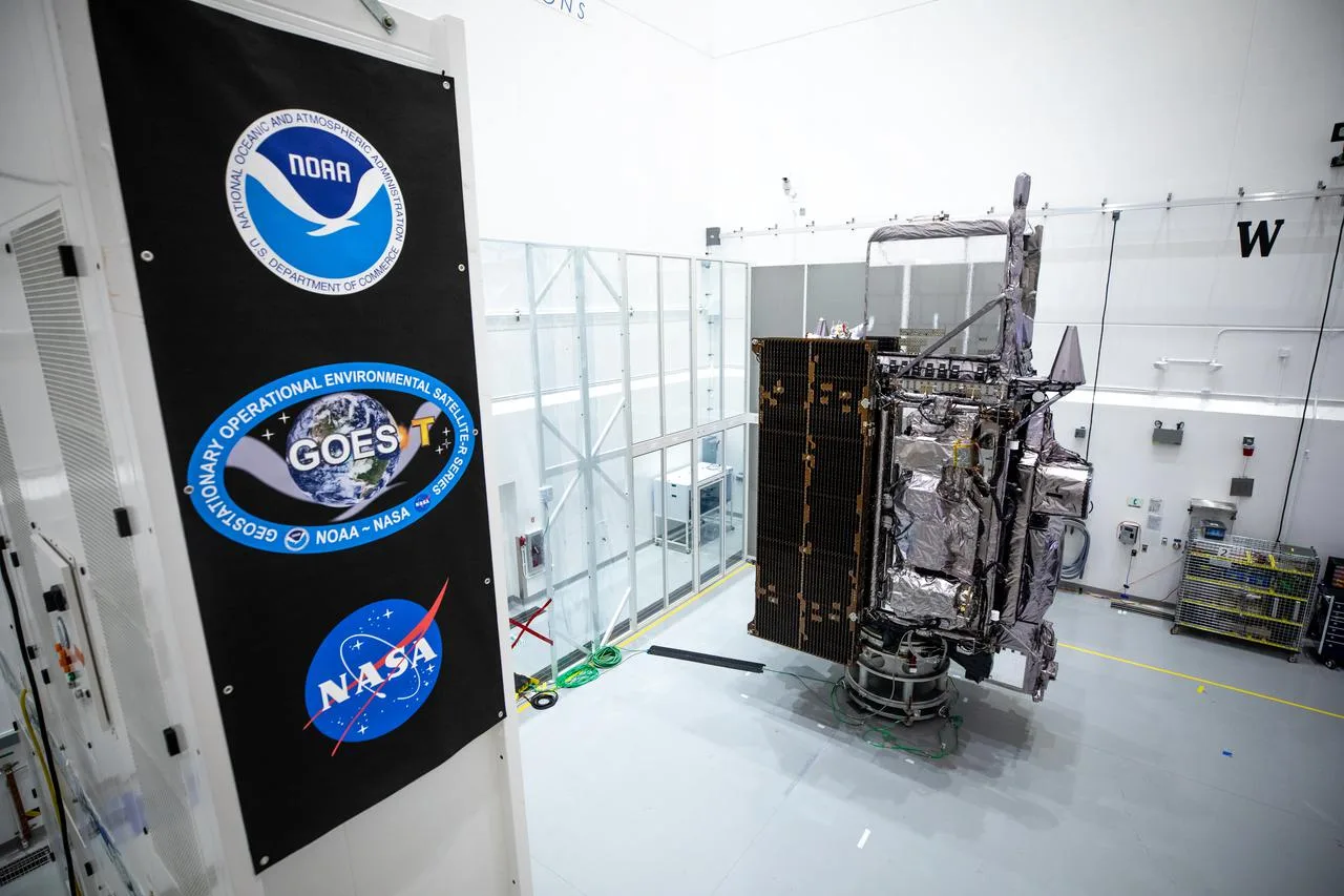 GOES-T satellite inside a cleanroom, wrapped in thermal insulation with solar panels extended and a nearby panel showing NOAA and NASA logos.