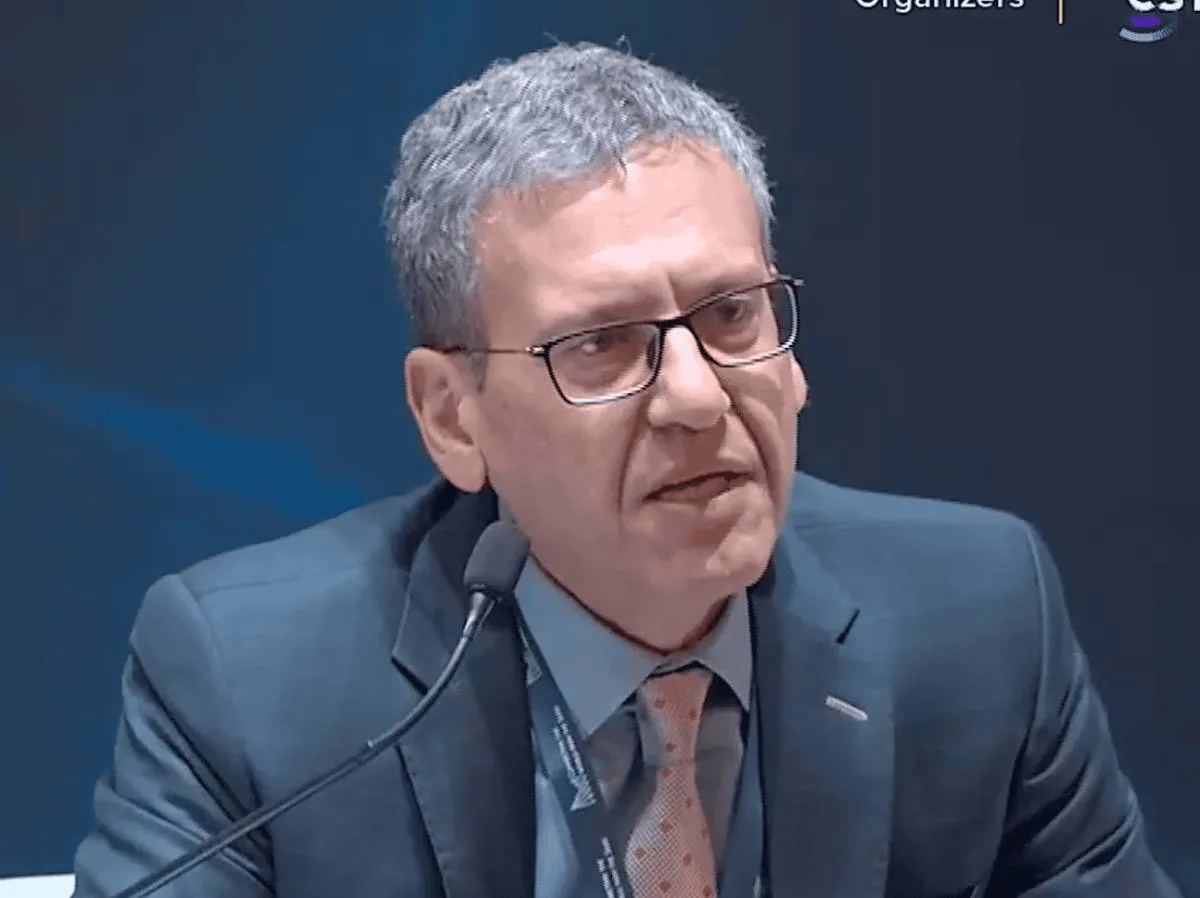 Hamid Akhavan speaks into a microphone at a formal event, wearing a suit and glasses.