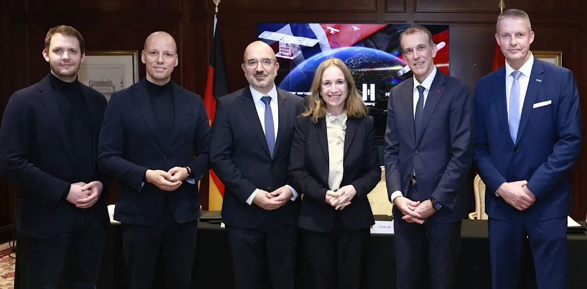 Six formally dressed officials stand side by side behind a table in front of a space-themed digital screen and a German flag, from left Daniel Metzler, Gundbert Scherf, Bernhard Kluttig, Laila Stenseng, Eirik Lie, and Dietmar Thelen.