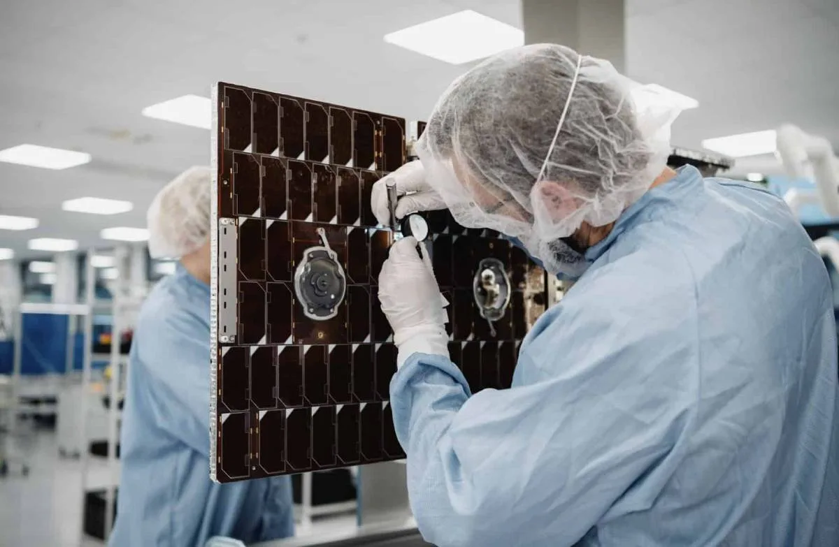 Technician in a cleanroom wearing a hairnet, face mask, gloves and blue lab coat inspecting a grid-pattern satellite solar panel array with a handheld tool.