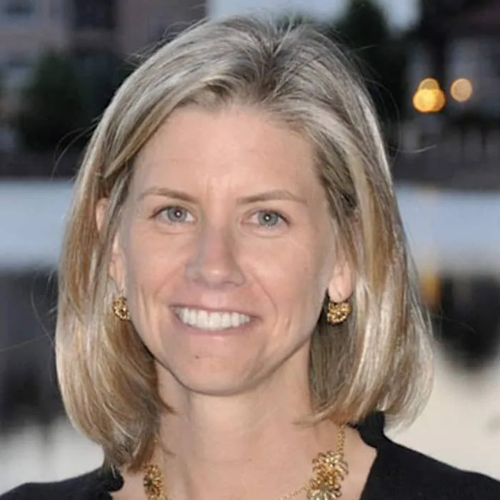 Head-and-shoulders portrait of Amy Mehlman, Lynk executive vice president and chief global affairs officer, with shoulder-length blonde hair wearing a black top and gold earrings and necklace against a softly blurred evening city background.
