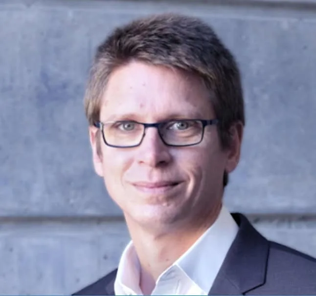 Daniel Troendle, wearing glasses and a suit, smiling slightly against a gray textured background.
