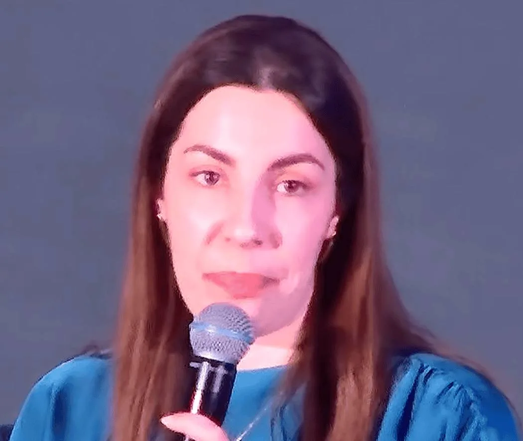 Theodora Liameti holds a microphone while speaking against a plain background.