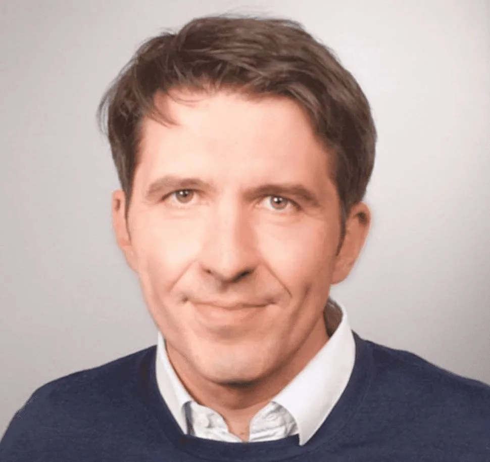 A headshot of Charles Delfieux, a man with brown hair wearing a white collared shirt under a dark blue sweater, set against a plain light gray background.